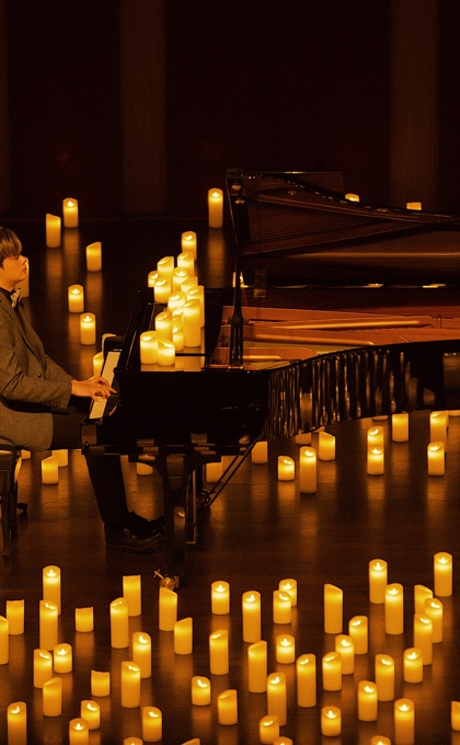 A side view of a man wearing a suit and playing the piano on a dark stage surrounded by hundreds of candles